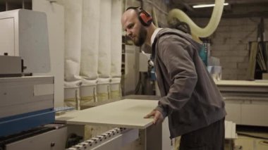 Mobilya atölyesindeki ahşap paneldeki kenar bantlama makinesini koruyan koruyucu kulaklık takan profesyonel bir marangoz.