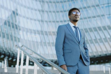 Kendinden emin iş adamı, güneşli bir günde modern cam binaya karşı duran takım elbiseli. Mesafeye bakarken yüzündeki kararlılığa bak.