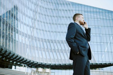 İş elbisesi giyen biri modern cam binanın dışında cep telefonuyla konuşuyor. Şehir ortamında profesyonel işlere odaklandığını gösteriyor.