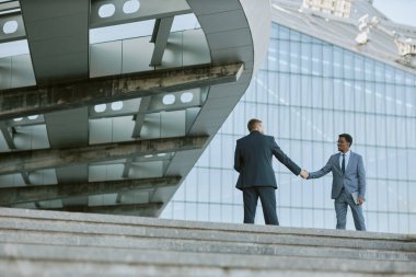Modern ofis binasının önünde el sıkışan iki işadamı cam cepheli profesyonel etkileşim ve çağdaş mimariyi yakalıyor.