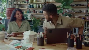 Genç adam ahşap masada kendisine dijital tablette oturma odasının modelini gösteren kadın tasarımcıyı selamlıyor.