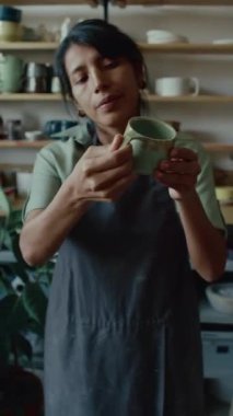 Siyah önlüklü esmer Latin zanaatkar kadının seramik bardak tutarken ve çömlekçi stüdyosunda incelerken dikey görüntüsü.