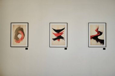 Galeri duvarında sergilenen modern soyut sanat eserlerinde siyah çerçeveli üç farklı parça dikkat çekiyor kırmızı ve siyah dinamik fırça darbeleri