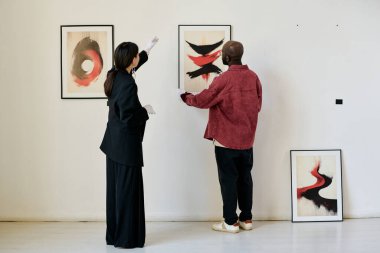 Beyaz bir galeri duvarındaki çerçeveli resimlere hayran olan ve tartışan iki kişi. Sanat eserleri çarpıcı bir görsel deneyim yaratan cesur renkler ve dinamik biçimlere sahiptir.