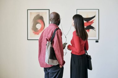 Sanat galerisinde iki kişi sohbet ederken beyaz bir duvarda çerçevelenmiş resimleri inceliyor. Görsel cazibeyi arttıran soyut biçimlerdeki çeşitli sanatsal ifadeler