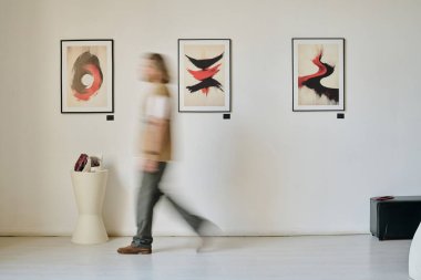 Bulanık bir figür, sanat galerisinde yürüyor. Duvarda hareketini gösteren soyut resimler sergiliyor. Sanat eserleri dinamik fırça darbeleri ve canlı karşıtlıklar sergiliyor