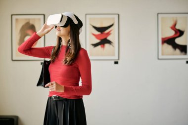 Modern sanat sergisinde sanal gerçeklik kulaklığı takan bir kadın. Arkaplanda çerçeveli sanat eserleri ile dijital deneyimi ayakta tutmak ve keşfetmek.