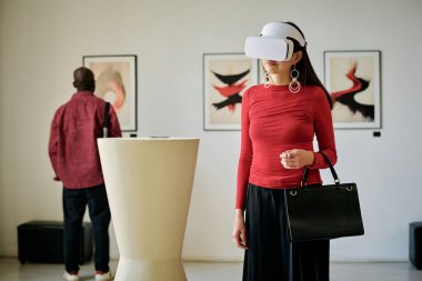 Sanal gerçeklik kulaklığı takan bir kadın çerçevelenmiş sanat eserlerinin yanında dururken bir sanat galerisini keşfediyor. Arka planda resimleri izleyen başka bir kişi