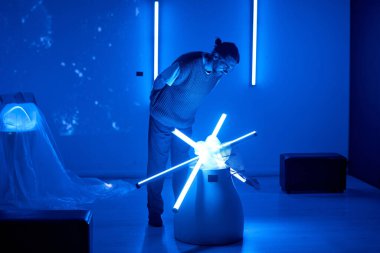 Mavi renkli odada aydınlatılmış silindirik desenli mavi neon tüpleri içeren sanatsal kurulumu gözlemleyen adam gelecekteki atmosferi yaratıyor.