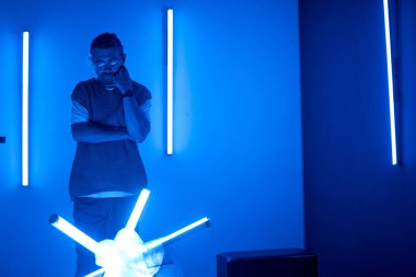 Neon ışıkları ve fütüristik dekor ile çevrili mavi ışıklı odada duran bir adam, düşünceli ve kendini çevresine kaptırmış görünüyor.