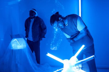 Mavi ışıklı bir odada gelişmiş teknolojiyle etkileşen iki birey, büyüleyici ve fütüristik bir ortam yaratıyor. Neon ışıklarının gölgeleri bilim kurgu atmosferini güçlendirir