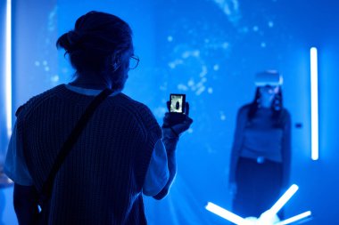 Akıllı telefon ile sanal gerçeklik deneyimini yakalayan kişi, mavi ışıklandırılmış ortamda teknolojiyle etkileşimi vurguluyor