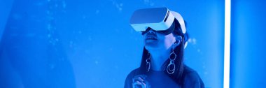 Woman wearing VR headset enjoying an immersive virtual reality experience, illuminated by a deep blue light. Large hoop earrings and long hair add to her futuristic look clipart