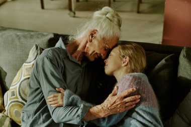 Rahat bir oturma odasında kanepede otururken küçük bir çocuğu kucaklayan yaşlı bir kadın. Nesil bağını ve sevgi dolu ilişkiyi vurgulayan sıcak etkileşim