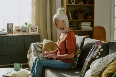 Oturma odasındaki rahat koltukta otururken dizüstü bilgisayar kullanan yaşlı kadın kitap rafları, bitkiler ve dekorasyonlarla sıcak ve davetkar bir ortam yaratıyor.