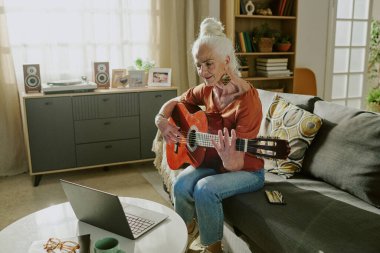 Rahat oturma odasında gitar çalan yaşlı bir kadın ve içeri ışık giriyor. Sehpada dizüstü bilgisayar, kitaplık ve arka planda saksı bitkileri aç