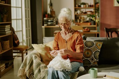 Kanepede oturan yaşlı kadın kitap ve çiçeklerle çevrili sıcak aydınlatılmış oturma odasında örgü örüyor.