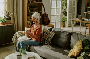 Kıdemli kadın rahat bir kanepede otururken örgü örüyor, güzel ışıklandırılmış oturma odasında çiçekler ve gitarla kitap rafında, huzurlu anın tadını çıkarıyor.