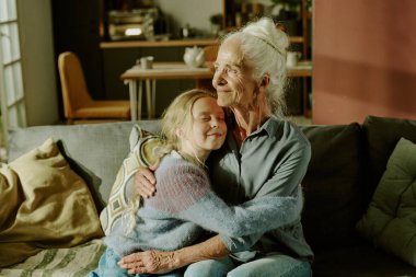 Senior woman with white hair embracing young girl on couch in cozy living room filled with natural light, creating heartwarming moment of affection and family bond clipart