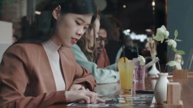 Genç Asyalı kadın yoğun bir restoranda kokteylle masada oturuyor ve menüdeki yemek resimlerine bakıyor, başka ne sipariş edeceğini seçiyor.