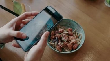 Kafedeki masada otururken sosyal medya için şehriyeli yemeğinin akıllı telefonuyla fotoğraf çeken bir kadının yüksek açılı görüntüsü.