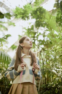 Sırt çantası takan, botanik bahçesinde yürüyen ve yeşil bitkilere bakan meraklı genç kızın dikey alçak açılı görüntüsü.