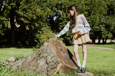 Teen schoolgirl spending time in botanical garden, touching huge old tree stump, long shot, copy space clipart