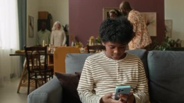 Oturma odasındaki koltukta otururken akıllı telefon kullanan genç çocuk, sonra aletleri kaldırdı ve öğle yemeği için dedelerini karşılamaya geldi.