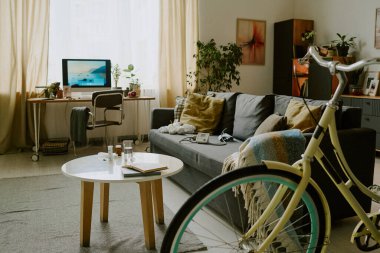 Koltuğun yanında bisiklet ve arka planda çalışma alanı olan rahat bir oturma odası. Saksı bitkileri ve boşluğa sıcaklık katan basit bir dekor.