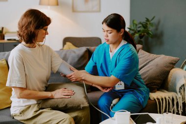Hemşire, rahat oturma odası ortamında, kanepede oturan yaşlı kadının tansiyonunu ölçüyor. İkisi de sakin ve odaklanmış bir sağlık prosedürü uyguluyor.