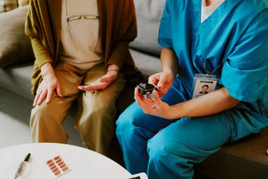 Mavi önlüklü hemşire yaşlılara ilaç veriyor. Sıcak oda ortamında koltukta oturuyor, yaşlı hastalara tıbbi bakım ve destek sağlıyor.