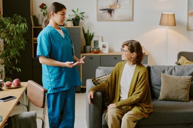 Hemşire, rahat bir yaşam alanında kanepede oturan yaşlı kadınla sağlık sorunlarını tartışıyor. Üst düzey kadın, sağlık uzmanlarının tavsiyelerini dikkatle dinliyor.