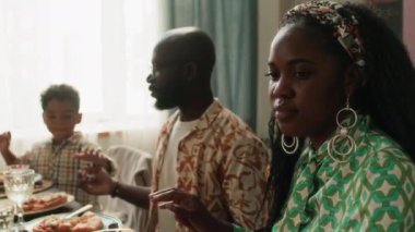 Yakın çekimde, Afrikalı Amerikalı bir kadının kocasıyla el ele tutuşup şenlikli aile yemeğinden önce dua ederken, arka planda küçük bir çocuk...