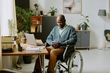 Tekerlekli sandalyedeki adam ev telefonuyla çalışırken etrafı kitaplar, bitkiler, dizüstü bilgisayarlar ve masalarla çevrili parlak, düzenli bir odada çalışıyor.