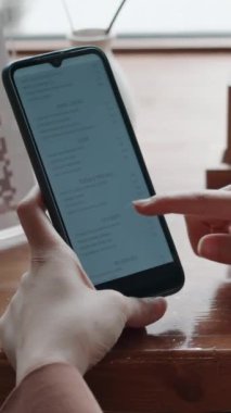 Akıllı telefonuyla menüyü kaydıran, kafede otururken ne ısmarlayacağını seçen, tanınmayan bir kadının dikey yakın çekimi.