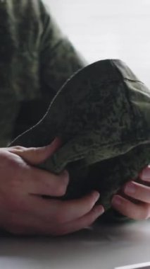 Kamuflaj üniformalı, elinde buruşuk bir şapkayla klinikteki doktor randevusundan önce endişelenen tanınmamış askerin dikey yakın çekimi.