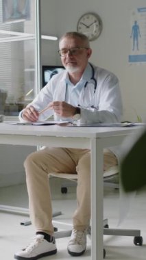 Tekerlekli sandalyedeki tanınmayan kıdemli askerin dikey fotoğrafı. Doktorla görüşüyor, muayenehanesinde oturuyor, randevu için.