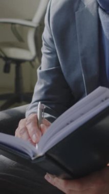 Not defterine yazarken ve danışmanlık ofisinde kameraya bakarken gri saçlı yaşlı erkek terapistin dikey olarak çekilmiş portresi.