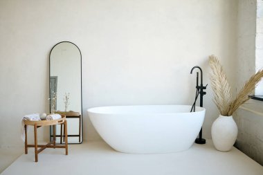 Kurumuş palmiye yapraklı büyük kil vazo ile minimalist banyonun arkasındaki siyah aynada dikdörtgen aynalı tahta masa arasında oval küvet.