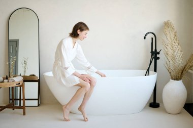 İpek bornoz giymiş genç bayan küvete oturmuş ve sıcaklığını ölçmek için suya dokunuyor.