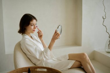 İpek bornoz giymiş, elleri yuvarlak aynalı ve ince çizgileri azaltmak için masaj yapan beyaz bir kadının yan görüntüsü.