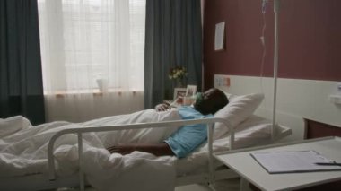 Oksijen maskeli mavi elbisesi olan Afro-Amerikalı bir adam, sırt üstü tek kişilik yatakta, parlak hastane koğuşunda enfüzyon standının yanında yatıyor.