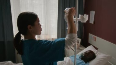 Tıbbi önlüklü genç bayan hemşire hastane yatağında uyurken oksijen maskesi takan yaşlı siyah adam için serum serumunu ayarlıyor.