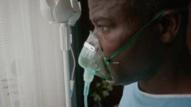 Açık mavi elbisesi ve oksijen maskesi takmış, şeffaf perdelerle hastane penceresinden dışarı bakan, elinde IV çubuğu olan yaşlı Afro-Amerikan bir adamın yakın çekimi.