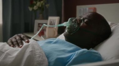 Hastane yatağında oksijen maskesi ve tüp takılı uyuyan, tıbbi giysili yaşlı Afrikalı Amerikalı bir adam.