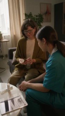 Dikey olarak, yaşlı bir kadın, reçeteli hapları hakkında tıbbi önlüklü genç hemşireye sorular soruyor.