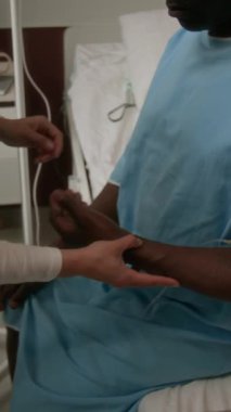 Önlüklü genç hemşirenin dikey fotoğrafı. Hastanenin koğuşunda oturan hastane önlüklü adamın serumunu kontrol ediyor.