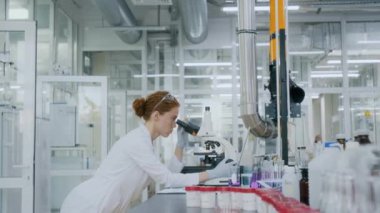 Beyaz önlüklü beyaz kadın laboratuarda çalışırken mikroskop kullanıyor ve gözlemlerini yazıyor.