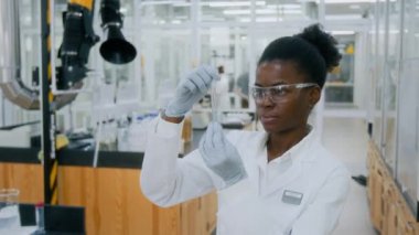 Elinde test tüpüyle kameraya bakan koruyucu gözlüklü Afrikalı Amerikalı genç kadın laboratuvar çalışanı portresi.
