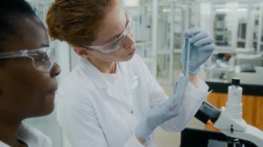 Dijital tabletli siyahi kadın kimyagerin laboratuvar ortamında test tüpünün içeriğine bakan Kafkasyalı iş arkadaşıyla yakın çekim görüntüsü.